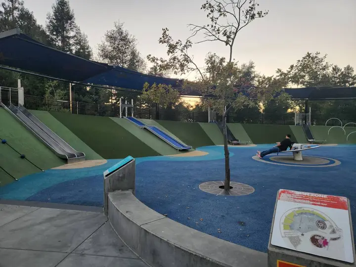 Turf hill with metal slides and a spinner on blue rubber at Magical Bridge Playground
